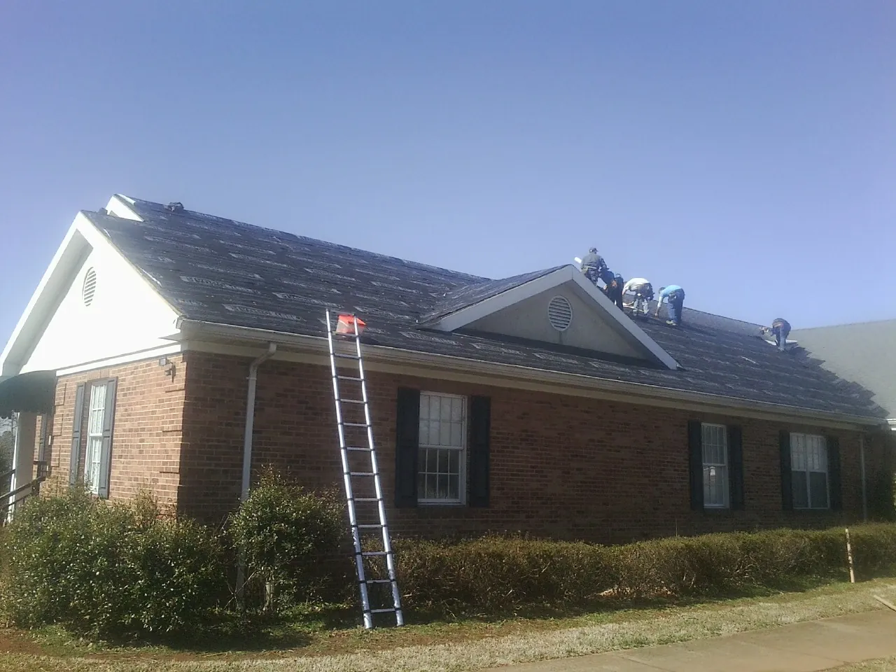 Crew tearing shingles off a brick ranch roof with a ladder set — Emergency Roof Repair work by our crew in Liberty Mills