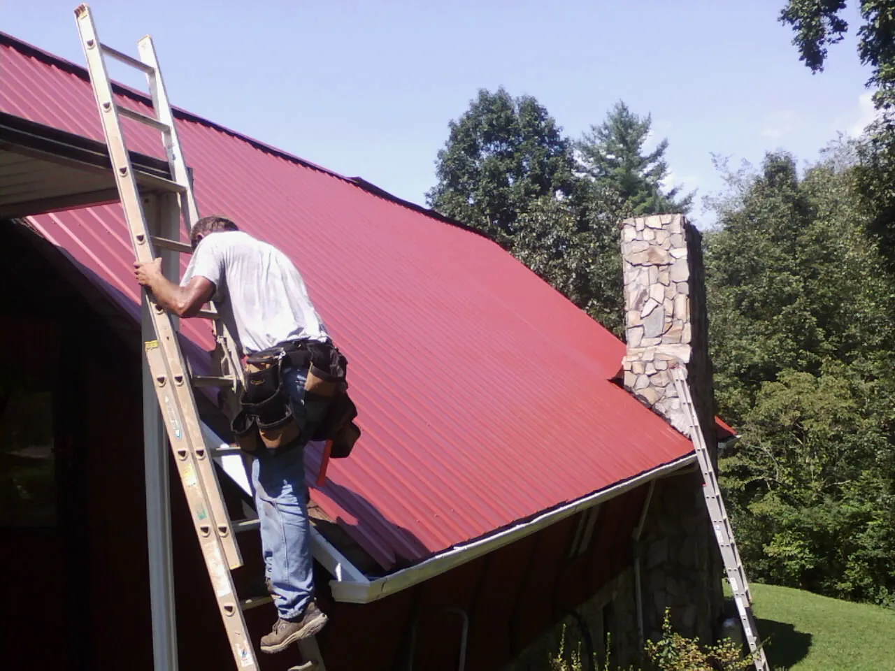 Roofer on a ladder beside a red metal roof and stone chimney — Soffit & Fascia Repair by Roofing Brothers in Liberty Mills